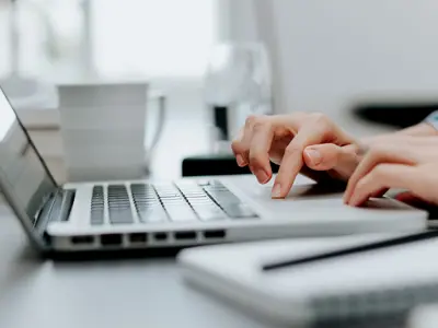 Close-up of hands working on a laptop with a pen and paper in the foreground.